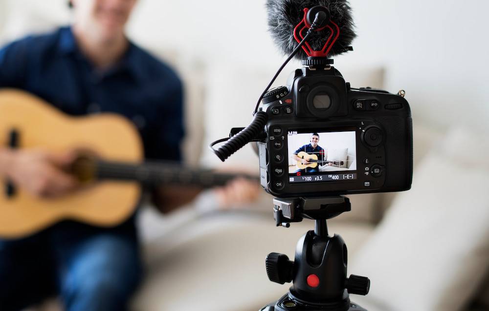 A camera is set up on a tri-pod to record a man while he practices guitar.